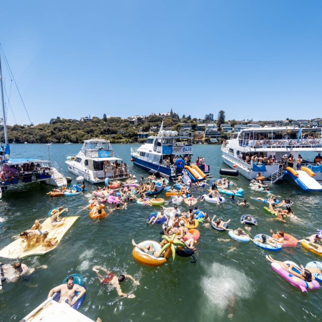 People are enjoying a sunny day on the water, swimming and floating on various inflatable tubes and rafts, surrounded by a group of anchored yachts. The clear sky and distant shoreline create a lively and festive atmosphere. A logo in the corner reads "The Yacht Social Club.