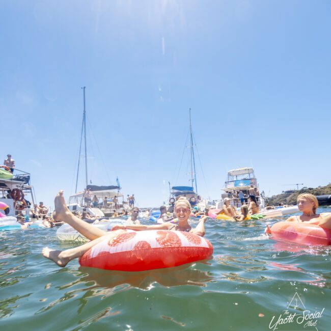 A joyful group of people in swimsuits float on inflatable tubes in clear, sunny waters. Several boats and yachts are anchored in the background under a picturesque blue sky. The festive mood is palpable, encapsulating the essence of Yacht Social Club, whose logo graces the bottom right corner.