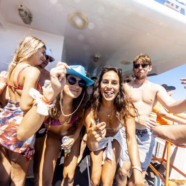 A group of people on a yacht enjoying a sunny day. They are wearing swimsuits, sunglasses, and hats while smiling and posing for the camera. The background shows a clear blue sky and calm water. The atmosphere is lively and festive.