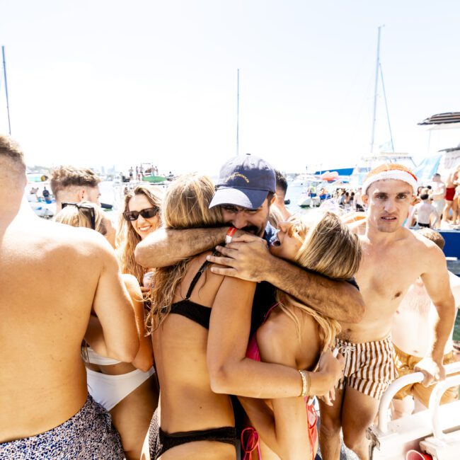 A group of friends in swimsuits gather closely together for a hug on a sunny day. In the background, boats and happy beachgoers enjoy the weather. The scene is festive, with a sense of camaraderie and celebration by the waterfront.