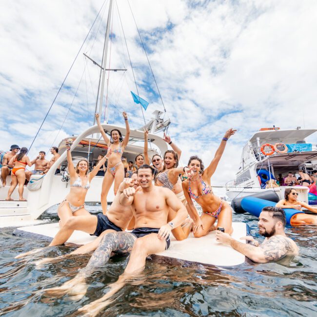 A joyful group enjoys a day on a luxurious yacht, some seated on its edge while others stand. The partly cloudy sky enhances the lively atmosphere as people in swimwear relax, with some taking a refreshing dip in the water beside it.