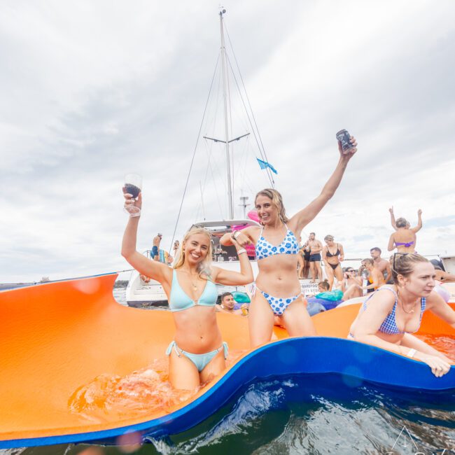 A group enjoys a day on the water, with two women in the foreground holding drinks and posing on an orange and blue floating mat. Wearing blue and white bikinis, they smile as a boat with a mast sails nearby. The atmosphere is cheerful and lively.