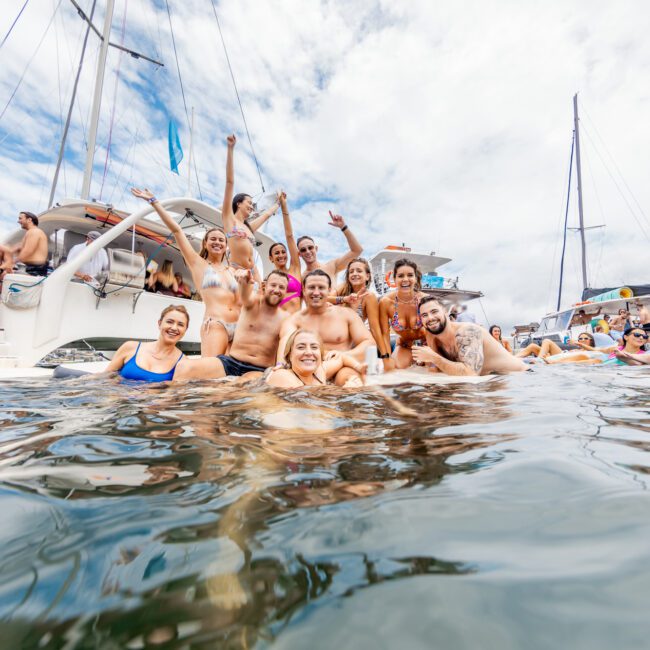 A group of people in swimsuits are smiling and posing together in the water with sailboats in the background. The weather is partly cloudy, and the scene exudes a festive and joyful atmosphere reminiscent of a summer celebration. The logo "Yacht Social Club" is visible in the bottom right corner.