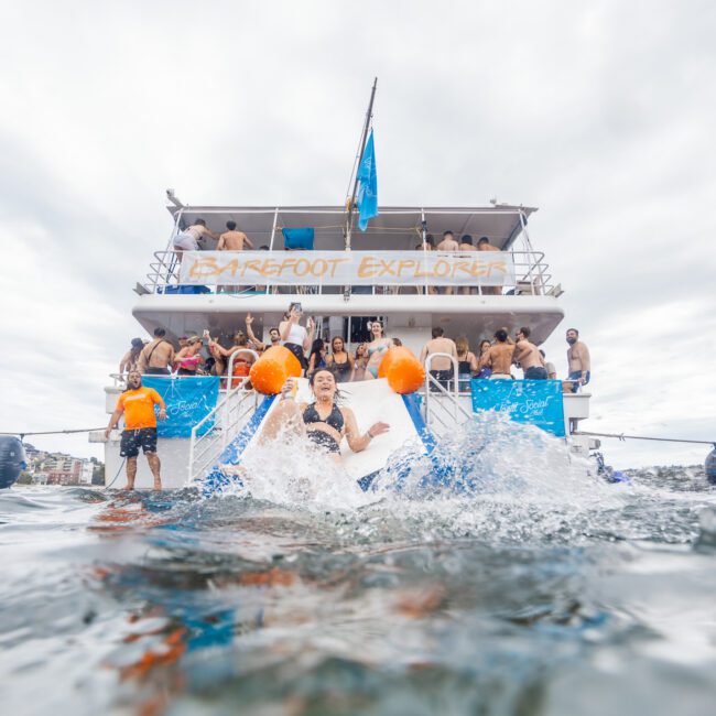 A group of people enjoys a day on a double-decker boat named "Barefoot Explorer." Some are on the upper deck, while a woman in a black swimsuit slides off an orange slide into the slightly choppy water. Despite the cloudy sky, everyone seems to be having fun paddleboarding nearby.