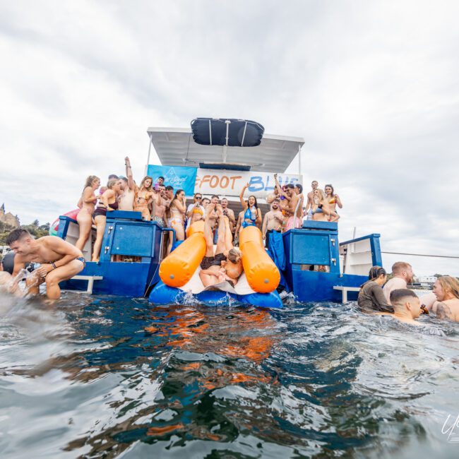 A group of people in colorful swimsuits are enjoying a floating bar on the water, with some sliding down orange slides into the sea. The scene is lively, with individuals swimming around the bar and standing on the platform sipping refreshing drinks under an overcast sky.