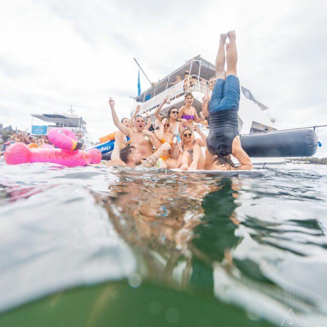 A group of people are partying on a boat and in the water, enjoying a sunny day in swimwear. One person is captured mid-dive into the water headfirst, while colorful inflatable pool toys float nearby. A majestic yacht anchored in the background adds to the festive scene.