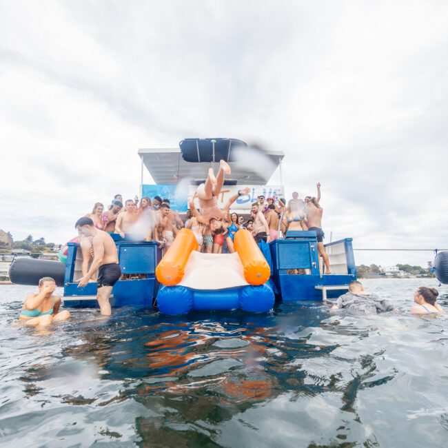 A group of people enjoying a party on a floating platform with a small waterslide, anchored on a lake or river. Several individuals are standing, sitting, or entering the water, while others watch from the upper deck. Kayaks are scattered around and trees and buildings are visible in the background.