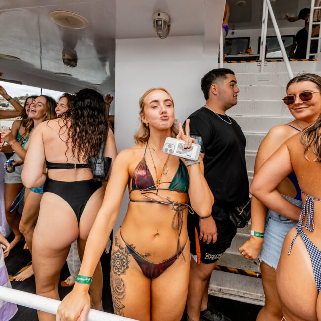 A group of people enjoying a lively party on a luxurious yacht. In the center, a woman wearing a bikini holds a drink and flashes a peace sign. Other guests around her engage in conversation and savor the event, while the yacht's elegant stairs and structure create an inviting backdrop.