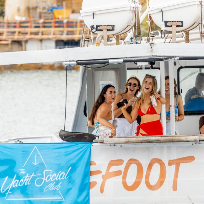 A group of people enjoying themselves on the deck of a boat labeled "SEFOOT." The boat is part of "The Yacht Social Club," according to a banner on the side. Several women are posing for the camera, smiling and laughing, with water and modern architecture in the background.