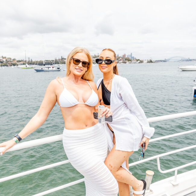 Two women in stylish swimwear and cover-ups lean against a railing on a boat, both wearing sunglasses and smiling. One holds a colorful can, while the other has her phone. The background features a picturesque body of water with boats and buildings in the distance under an overcast sky.