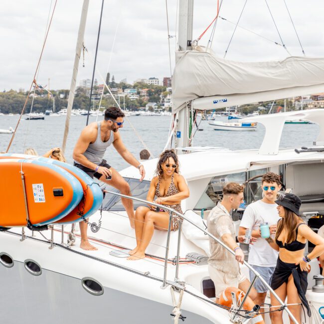 A group of people enjoying a sunny day on a yacht. Some are sitting on the deck, while others are standing and conversing. One man in sunglasses and a tank top leans on the railing. The background features a calm body of water and a shoreline with buildings, highlighting The Yacht Social Club Sydney Boat Hire experience.