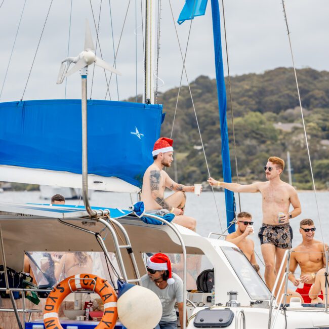 A group of men is enjoying a sunny day on a white sailboat named "Catalyst Sydney." One man, wearing a Santa hat and holding a drink, sits on the deck. Several others are relaxing, some shirtless and one also in a Santa hat. Lush hills and trees create a picturesque background.