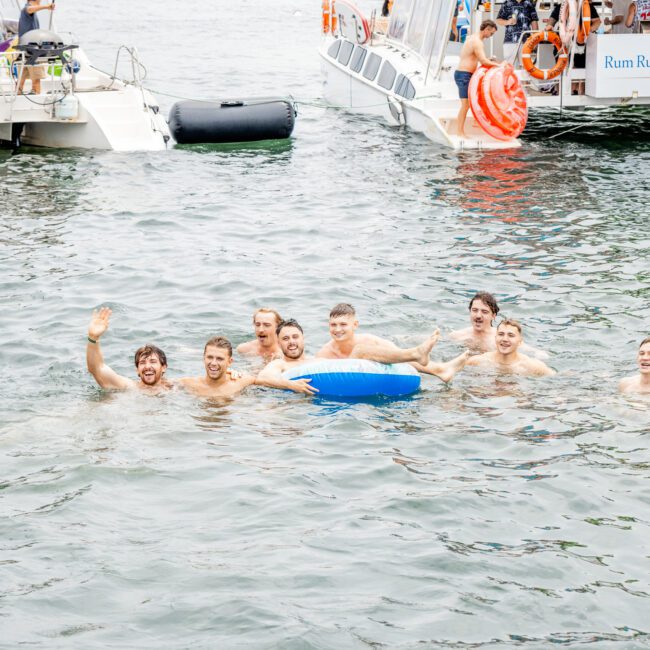 A group of eight people is swimming in the water, some of them holding a blue tube. They are smiling and waving towards the camera. In the background, two boats with people onboard and life-saving equipment are anchored nearby.