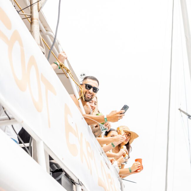 A group of people on a boat enjoying themselves. One man wearing sunglasses is smiling and holding a camera, while a woman behind him takes a photo with her phone. Another woman in a straw hat sips an orange drink under clear skies. A lively banner flutters in the background.