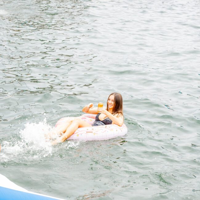 A person is relaxing on an inflatable ring in the water, holding a drink, with a luxury yacht and other people visible in the background. The serene water enhances the leisurely moment as they soak up the tranquility.