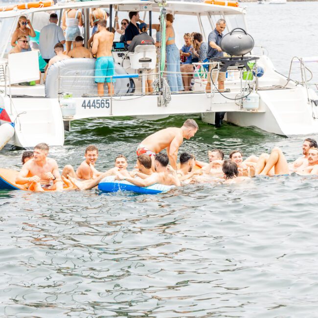 A group of friends enjoys a sunny day, gathering on a floating inflatable raft near a docked boat. Some stay aboard while others relax on the raft, creating a lively summer scene by the water's edge.