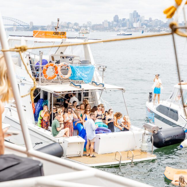 A group of people enjoying a lively party on a boat in a harbor, with the city skyline visible in the background. The boat is decorated with colorful banners, and there is another smaller vessel nearby. The day appears to be bright and slightly overcast, creating a picturesque setting for celebration.