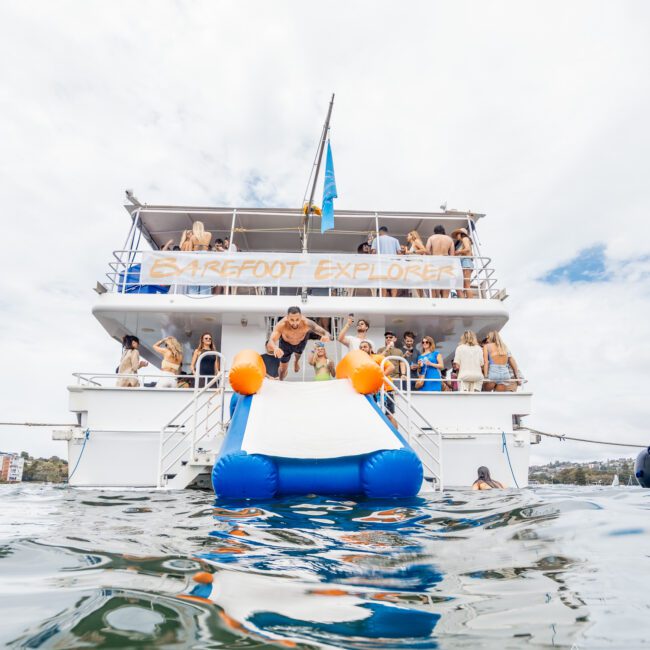 A lively scene of a two-deck boat named "Barefoot Explorer," brimming with people enjoying festivities. In the foreground, a vibrant water trampoline floats in the blue and orange water, with a person mid-jump from the boat towards it, surrounded by captivated onlookers.