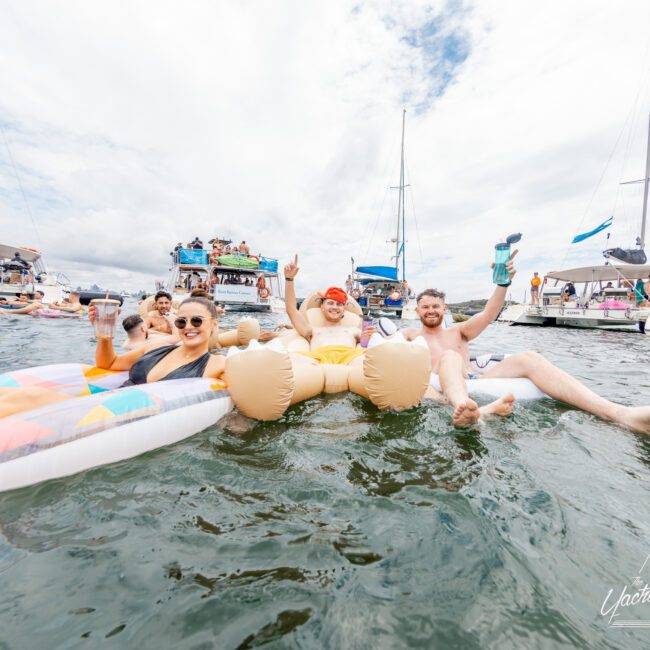 Three people are floating on inflatable rafts in the water, smiling and holding drinks. Several boats are in the background, with people also enjoying the water under a cloudy sky. An inflatable Santa Claus is prominently featured on one of the rafts, adding a festive touch to the scene.