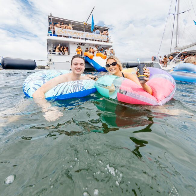 Two people relax on vibrant inflatable floats in the water near a party boat teeming with excitement. The sky is partly cloudy, and the boat's deck showcases a banner reading "Stepfoot Express." The atmosphere is lively and festive.