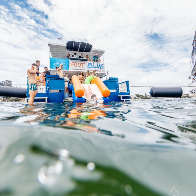 People are enjoying a sunny day on a blue boat named "Foot Blue," featuring a slide into the water. Some are standing on the boat, while others are in the water. The logo "The Yacht Social Club" is visible in the corner, with a partly cloudy sky as the backdrop—truly an invigorating scene.