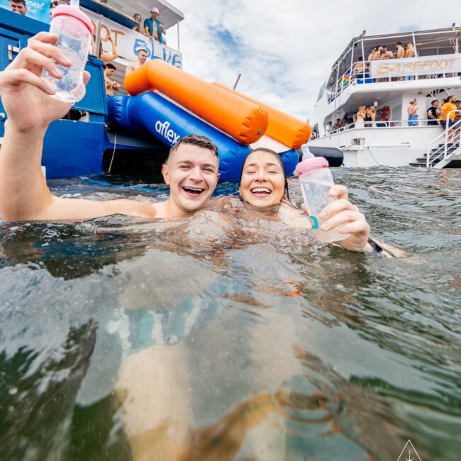 A man and woman are smiling and holding bottles while standing in the water near a blue and white boat with inflatables. Other people are on the boat, with another vessel in the background. The scene suggests a festive and social gathering on the water, reminiscent of a Yacht Social Club event.