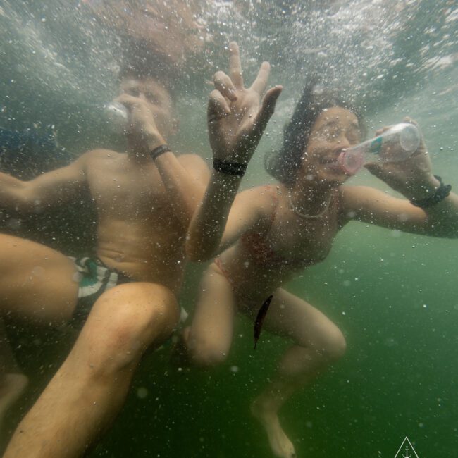 Two individuals are underwater, sipping from cans and cups as they swim. Bubbles and water movement surround them, highlighting their active motion. The person on the right flashes a peace sign with their hand. A watermark in the bottom right reads "The Yacht Social Club.