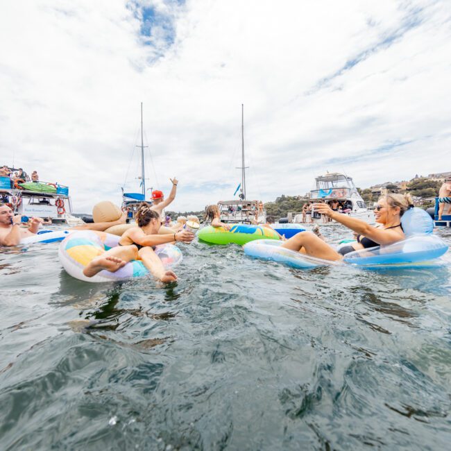 A group of people floating on inflatable rings in a body of water, surrounded by boats. The sky is cloudy, and a few people are taking photos while others are chatting and relaxing. The slightly rippling water adds to the lively atmosphere, creating an idyllic scene for everyone.