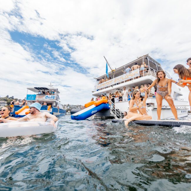 A lively scene with people enjoying a sunny day on the water. Some are on inflatable rafts and floating toys, while others are on a dock and boats. One boat features a slide, with skyscrapers towering in the background under a partly cloudy sky.