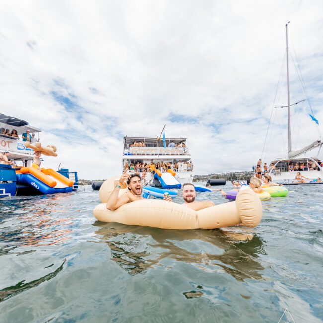 Two people float on an inflatable banana in the water at a crowded, festive boat party. Surrounding them are several boats, including a large double-deck yacht filled with people enjoying the lively atmosphere. The sky is cloudy, and other partygoers can be seen having fun on various inflatables and stand-up paddleboards.