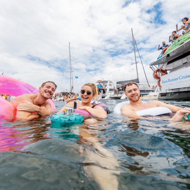 Three friends are floating in a body of water, each with inflatable rings. Two men flank a woman who enjoys drinks on a floating tray. Various yachts dot the background, with people aboard enjoying the festive and relaxed atmosphere.