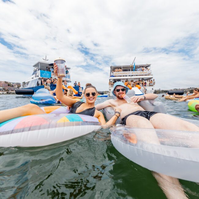 People float on pool inflatables in a lake, raising drinks and smiling. Two yachts with more people are in the background. The sky is partly cloudy, and the shoreline with houses is visible. The atmosphere is festive and lively. Text at the bottom reads "Yacht Social Club.