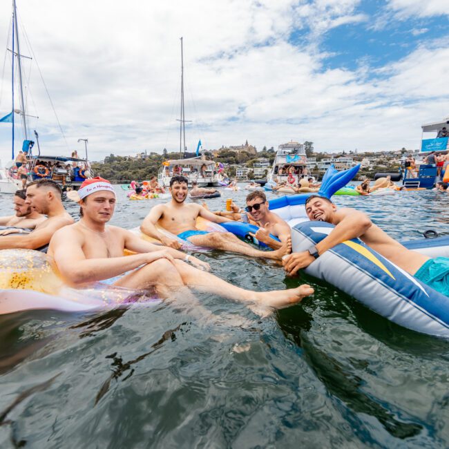 A group of people float and relax on inflatables in the water near a boat with a "Yacht Social Club" sign. Other boats and kayaks are visible around them, under an overcast sky, creating a lively, social atmosphere.