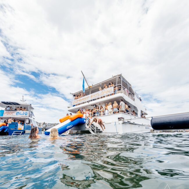 A large double-decked yacht is anchored on a sunny day, surrounded by people swimming and floating in the water nearby. An inflatable slide is attached to the back of the yacht, with people on the deck enjoying the scene. Another boat with colorful sails is visible to the left.