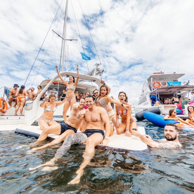 A group of friends enjoying a sunny day on a yacht, some swimming in the clear water and others relaxing on the deck. They appear to be having fun, posing for the camera with smiles and energetic gestures. Nearby, another boat with additional guests enhances the lively and festive atmosphere.