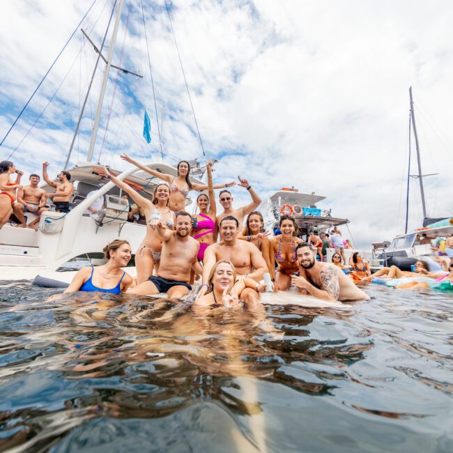 A large group of people is gathered around a few boats, enjoying a day on the water. Some are swimming, while others sit or stand on the boats, all smiling and appearing joyful. The sky is partly cloudy, and the scene is festive. A Cotton Social logo adds to the lively atmosphere.