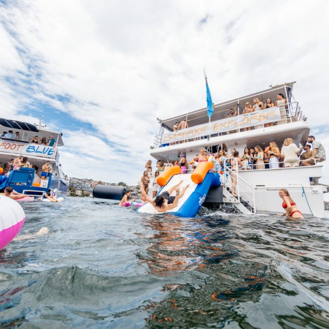 A lively scene with people enjoying a boat party on the water. Two boats are anchored close together, with people swimming and using colorful inflatables. One boat features a fun water slide, and many participants are relaxing on the decks, soaking up the sunny day.