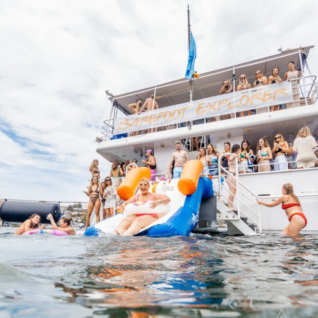 A group of friends enjoy a sunny day on a boat named "Barefoot Explorer." Some are on the deck, while others splash in the water using an inflatable slide. The water is calm, and the atmosphere is lively and fun. The Yacht Social Club logo is visible in the corner, adding to the sense of community.