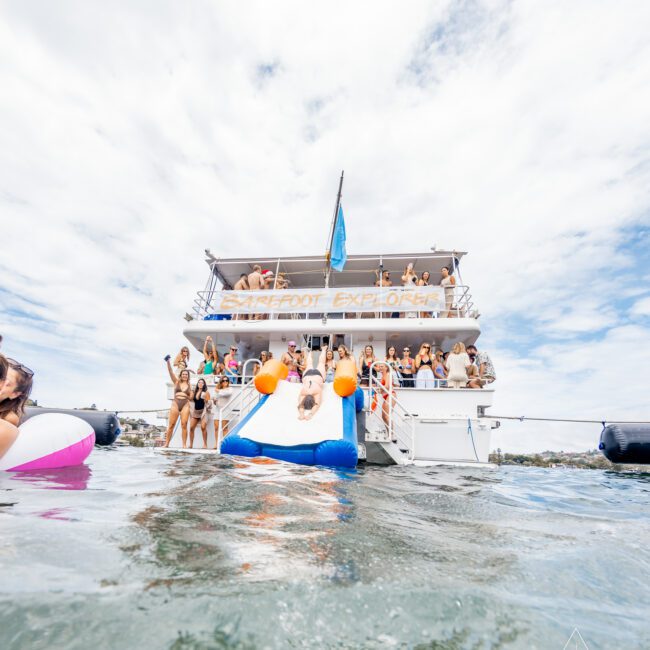 A large group of people enjoying a sun-drenched day on the two-deck boat "Barefoot Explorer." Some are lounging while others swim near the boat, accompanied by an inflatable float in the water. The scene is lively and festive against a backdrop of blue sky and calm water, creating an idyllic summer experience.