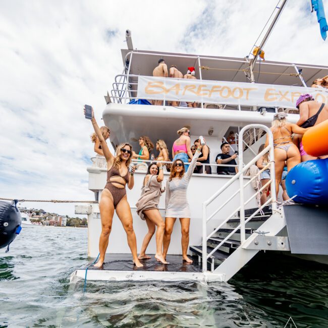 A lively group enjoys a boat party on the multi-deck vessel labeled "BAREFOOT EXPRESS." Three women in swimsuits and casual wear stand on a platform at the back of the boat, posing and having fun against the backdrop of a partly cloudy sky. The atmosphere is vibrant and carefree.