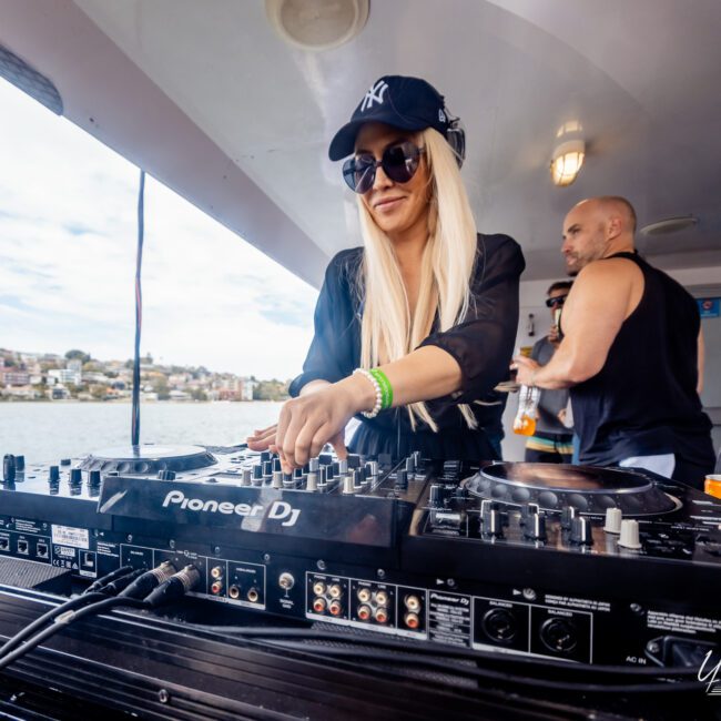 A person wearing sunglasses, a black outfit, and a baseball cap is DJing with a Pioneer DJ controller on a boat. Another person in the background is holding a drink and watching. The scene overlooks a pristine body of water with buildings visible in the distance, capturing an unforgettable vibe.