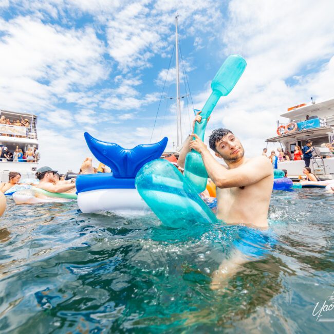 A man sits on a blue inflatable mermaid tail float in the water, rowing with a large blue paddle. Other people on various inflatables and yachts are in the background under a partly cloudy sky. Logo in the corner reads "Yacht Social Club".