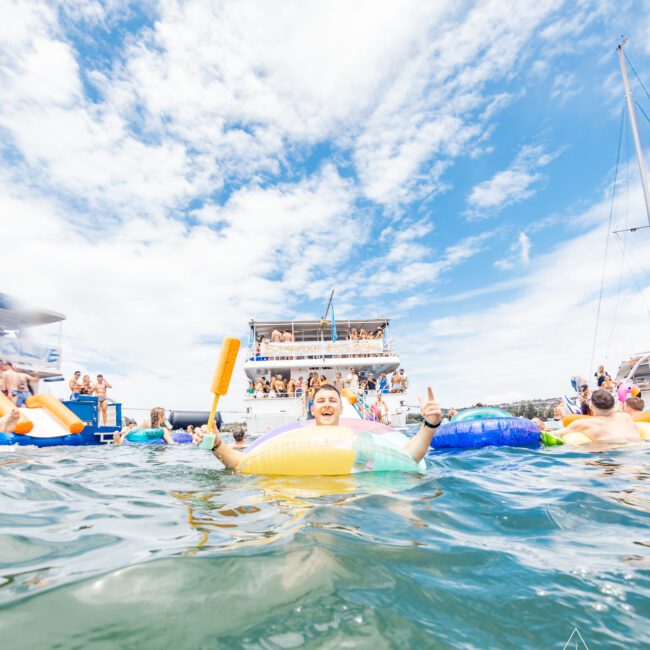 A man floats in the water with a colorful inflatable tube, holding an oar, and giving two thumbs up. In the background, a large boat is filled with people under a partly cloudy sky. More people are enjoying the water around him. The text "The Yacht Social Club" is seen in the bottom right corner.