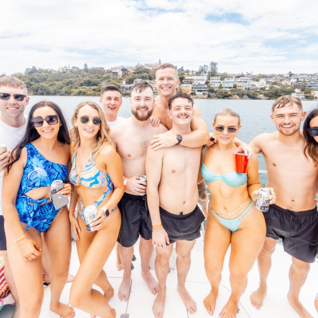 A cheerful group of nine, some in swimsuits, smile and pose for a photo on a boat. They stand close together, holding drinks, with water and a picturesque hilly area dotted with houses in the background. It is clearly a sunny day filled with fun.