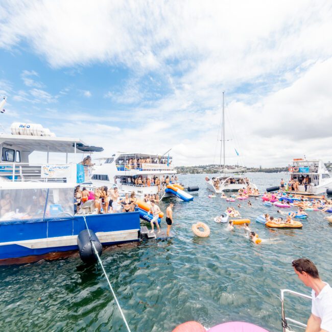 Boats and people gathered in a vibrant, blue-watered bay under a partly cloudy sky. Numerous colorful inflatables and swim rings float with people on them. One boat hosts socializing guests on its deck, contributing to the festive atmosphere.