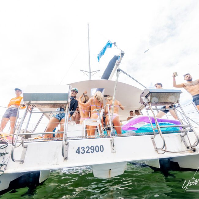 A group of people enjoy a lively day on a white boat, wearing swimsuits and summer attire. They are engaged in conversation and cheering with drinks. The boat has a license number 432900 and is surrounded by water with "Yacht Social Club" written in the bottom corner, enhancing the festive atmosphere.