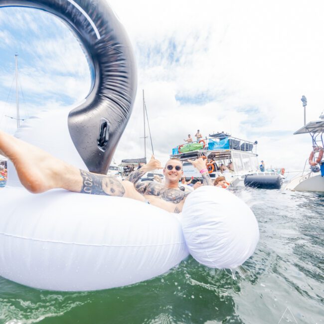 A person with sunglasses and tattoos enjoys lounging on a large inflatable swan in a body of water. Several boats and people can be seen in the background, indicating a lively social gathering, possibly an exciting beach party. The sky is partly cloudy.