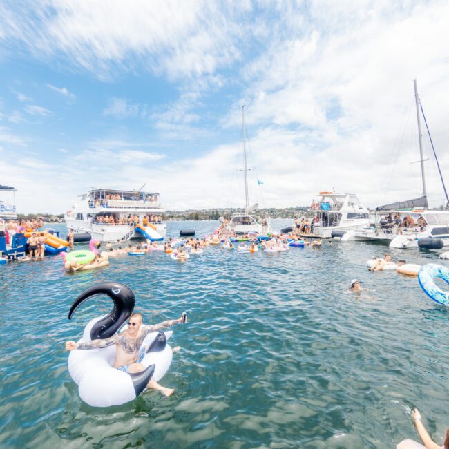 A lively scene on a sunny day shows people enjoying a boat party. Dozens of folks are in and around the water, partly on inflatable floats of different shapes and colors. Several boats are anchored nearby with more people. The sky is dotted with fluffy clouds, making for a pleasant atmosphere.