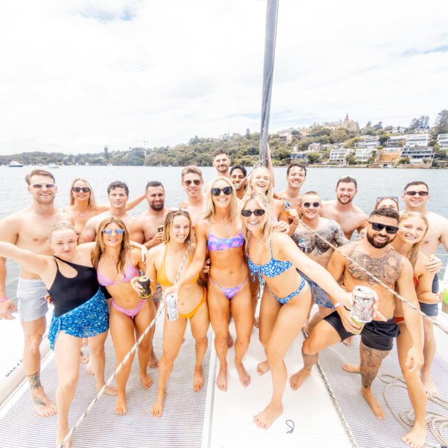 A large group of smiling people in swimsuits is posing on a sailboat in front of a coastal town. They are holding drinks and appear to be enjoying a sunny day on the water. The background features a hilly landscape with scattered houses.