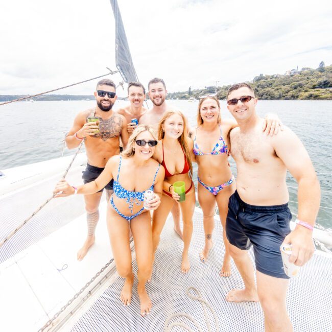 A group of seven people in swimwear are standing and smiling on the deck of a boat, holding drinks and enjoying a sunny day on the serene water. The background shows a calm body of water and a lush green shoreline.